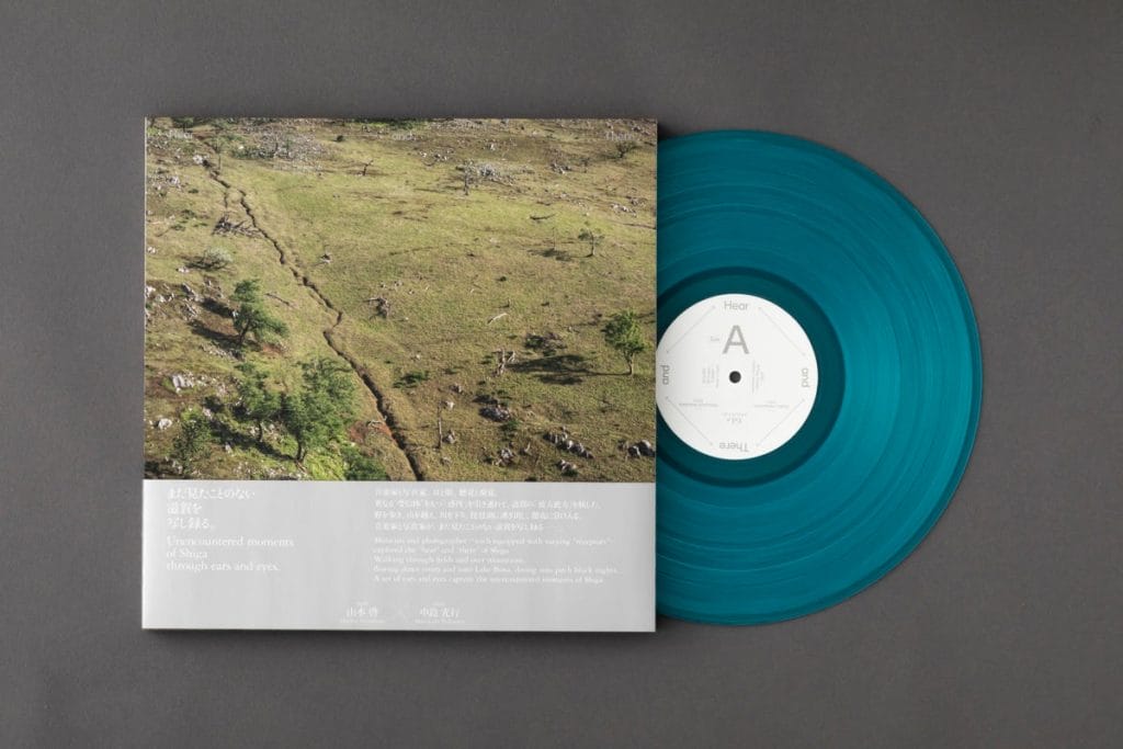 Teal vinyl LP partially slid out of a landscape album cover showing a grassy hillside with trees and rocks; bottom has white text blocks in English and Japanese.