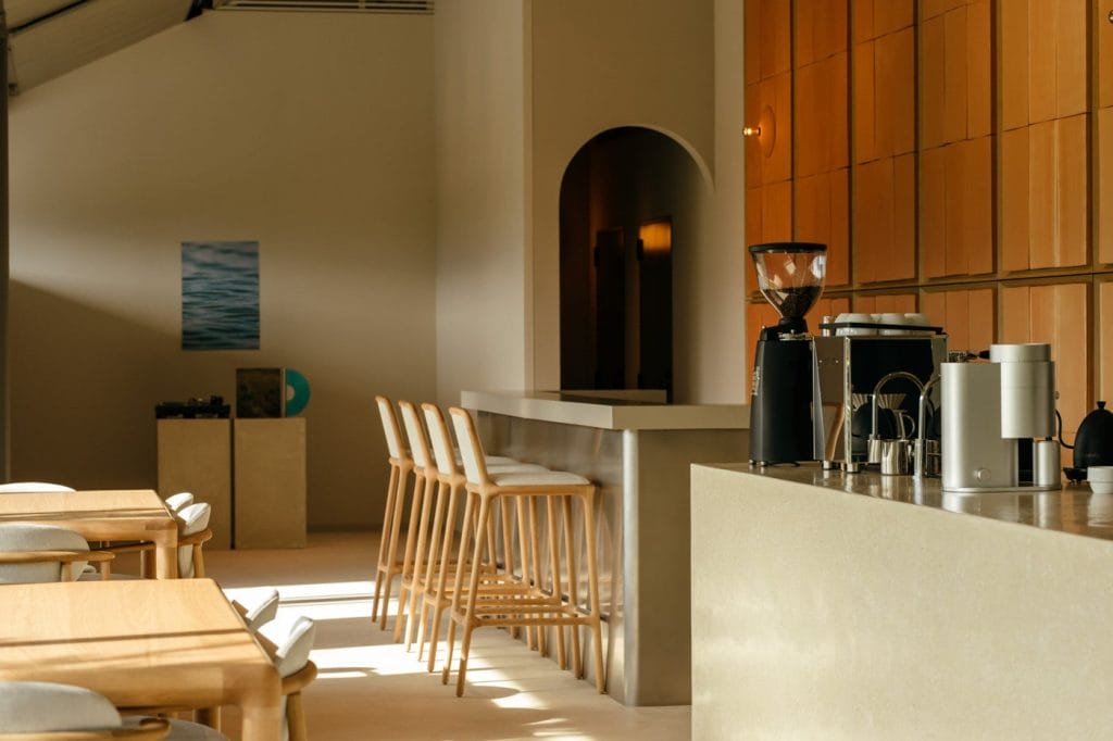 Modern cafe interior with a long light counter, row of wooden stools, and a coffee station on the right.