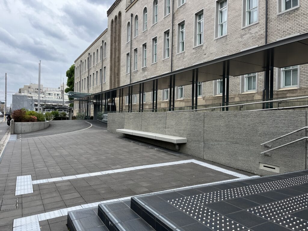 Exterior view of a long brick building with rows of tall rectangular windows and a covered walkway along the ground level. A sloped ramp and metal railings run beside it, with a tiled plaza in the foreground and cloudy sky above.