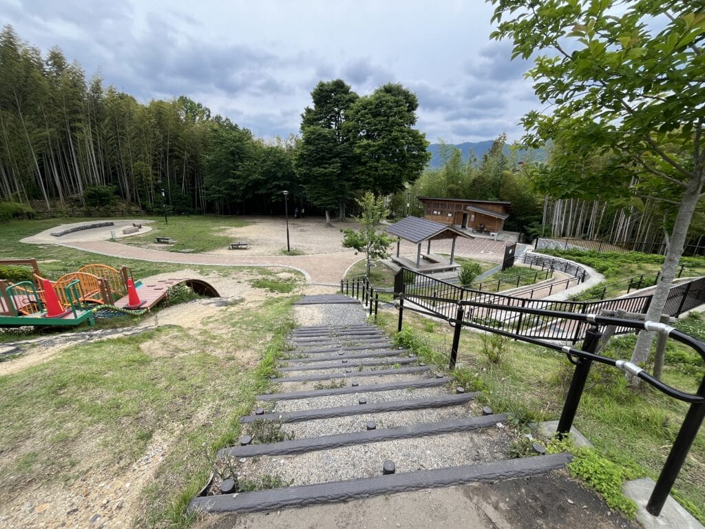 Descending stone steps with black railing into a park, playground equipment on the left, and trees under a cloudy sky in the background.