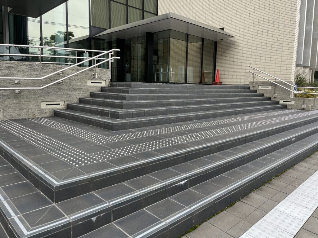 Wide, dark tiled stairs with metal handrails leading to a glass-front entrance of a modern building; a red cone sits near the doorway and tactile paving runs along the lower steps.