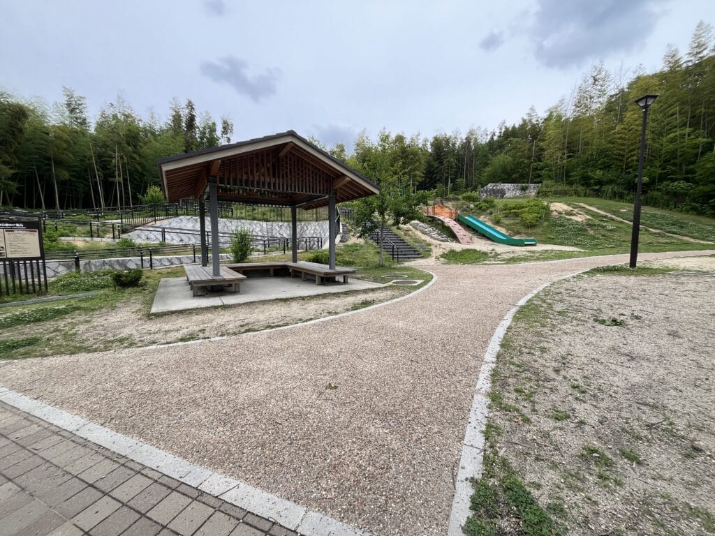 Park pavilion with a wooden roof, picnic benches, and a playground slide visible in the distance among curved gravel paths and trees nearby.