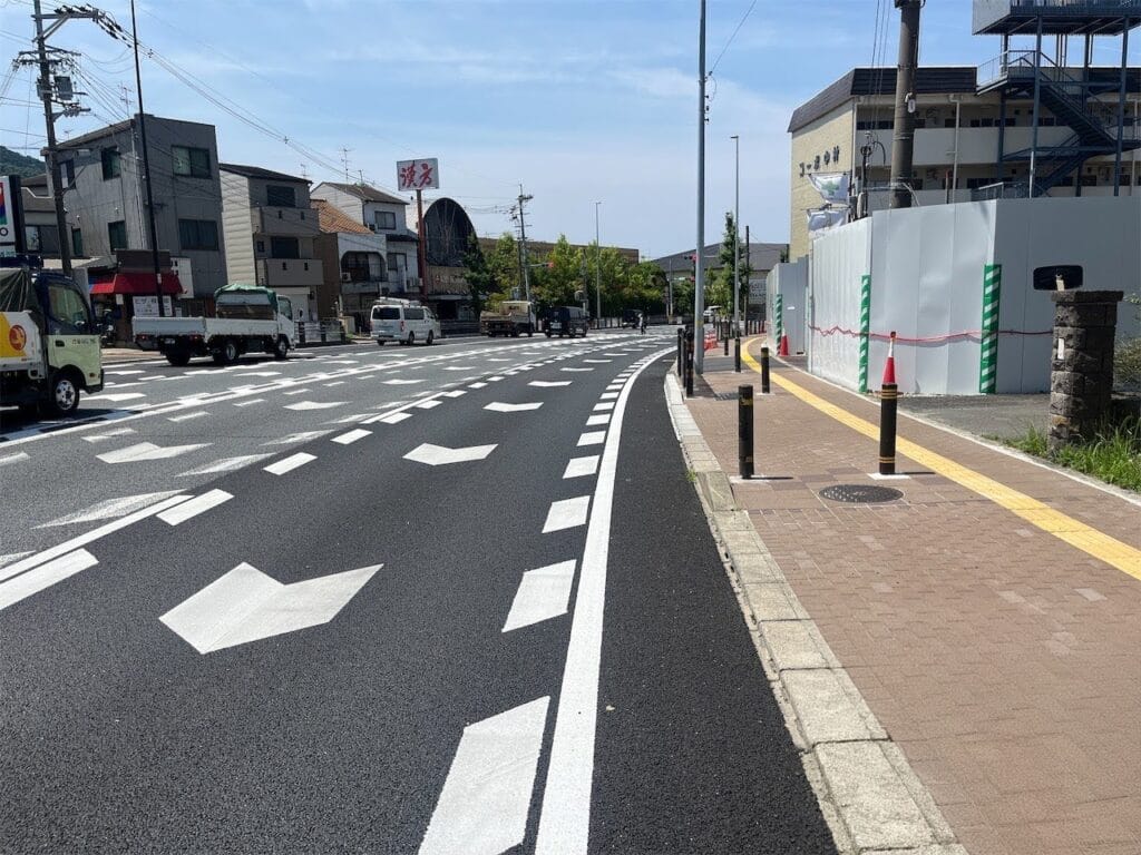 Urban street with multiple lanes and bold white lane markings, cars in the distance, and a sidewalk on the right under construction barriers