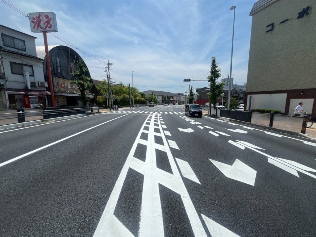 Wide multi-lane street in a Japanese city with bold white lane markings, a crosswalk ahead, and storefronts on both sides of the road.