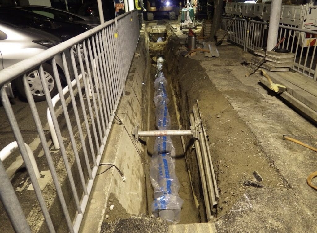 Nighttime construction scene: a deep trench along a sidewalk with temporary railing and construction equipment nearby.