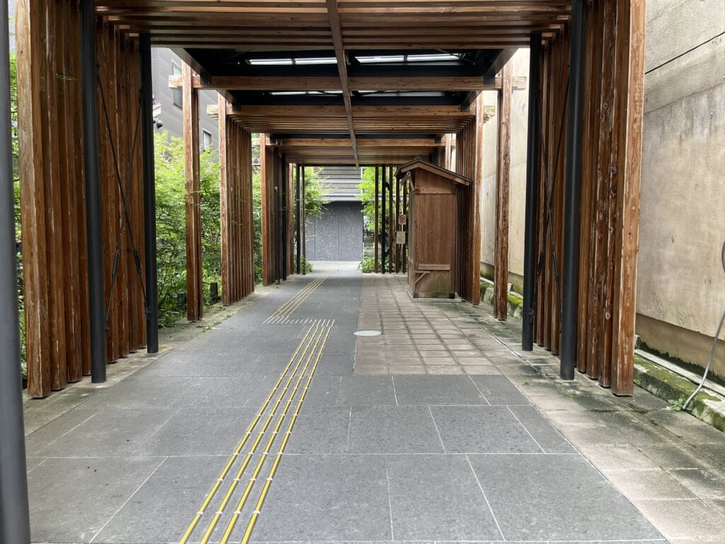 Wooden pillar walkway under a timber-framed cover, with yellow tactile guide lines along the path toward a small structure in the distance.