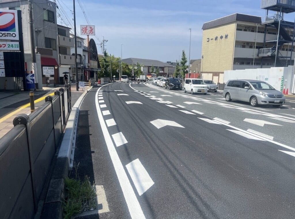 Wide city street in Japan with multiple lanes, white road markings, and cars traveling on the left under a blue sky.