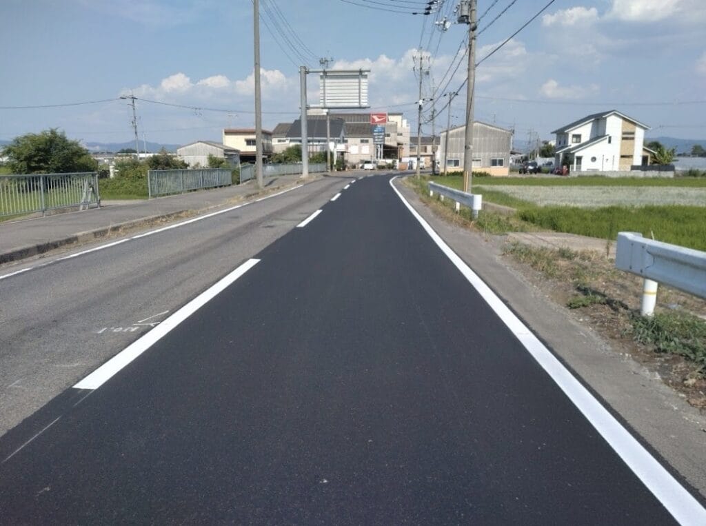 Newly paved two-lane road in a rural town, with white lane markings and guardrails on both sides.