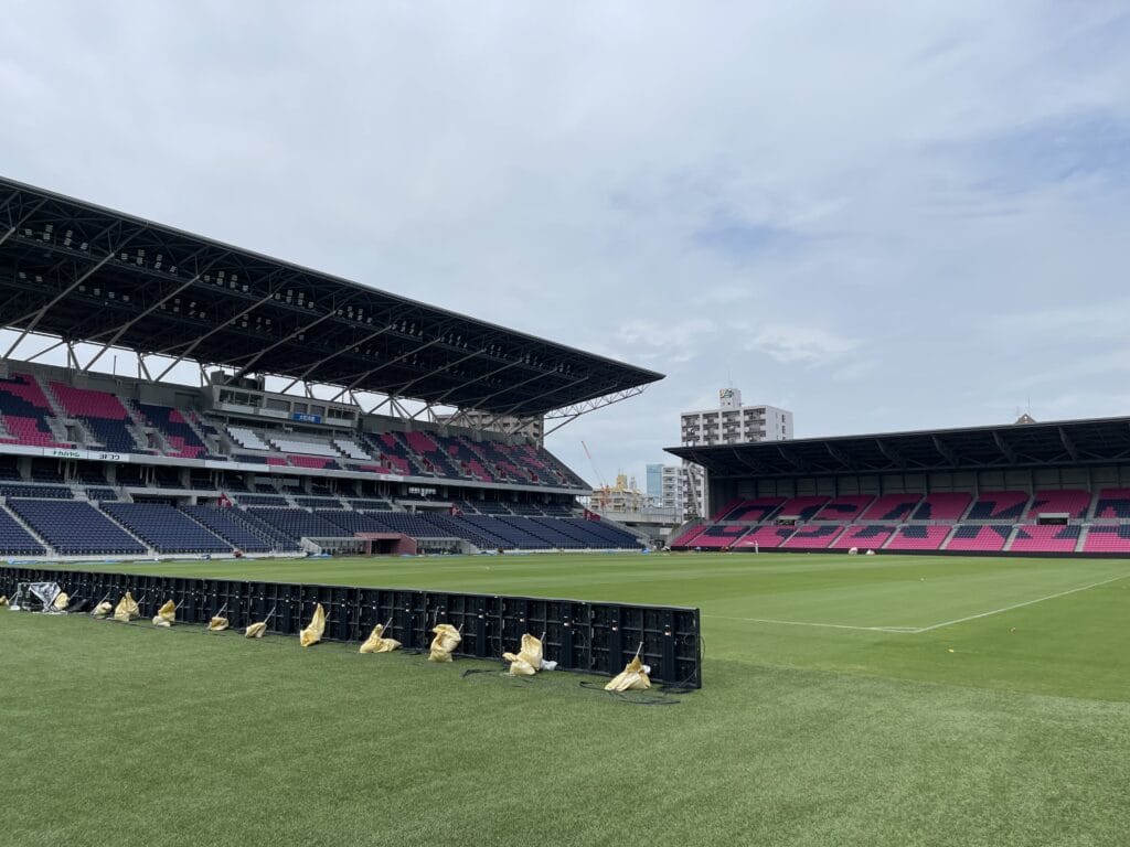 view of a sports stadium with tiered pink and blue seats under a large overhanging roof, construction barriers along the sideline.