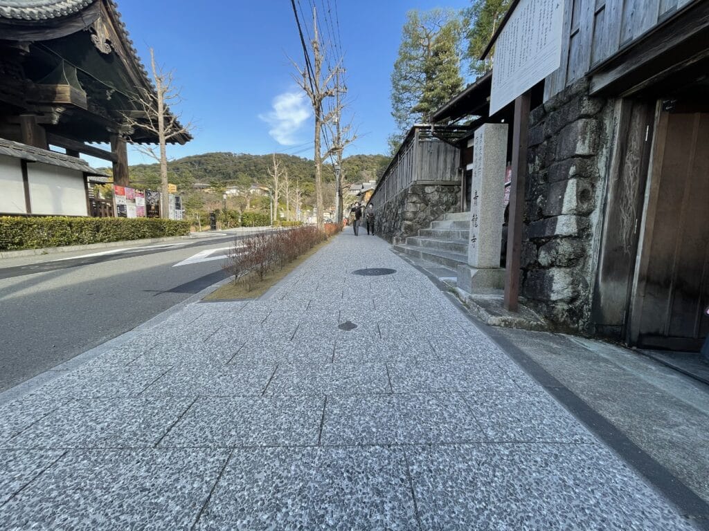 Stone-paved pedestrian street in a historic Japanese area, lined with wooden buildings, stone walls, and leafless trees; two people walk in the distance under a blue sky.