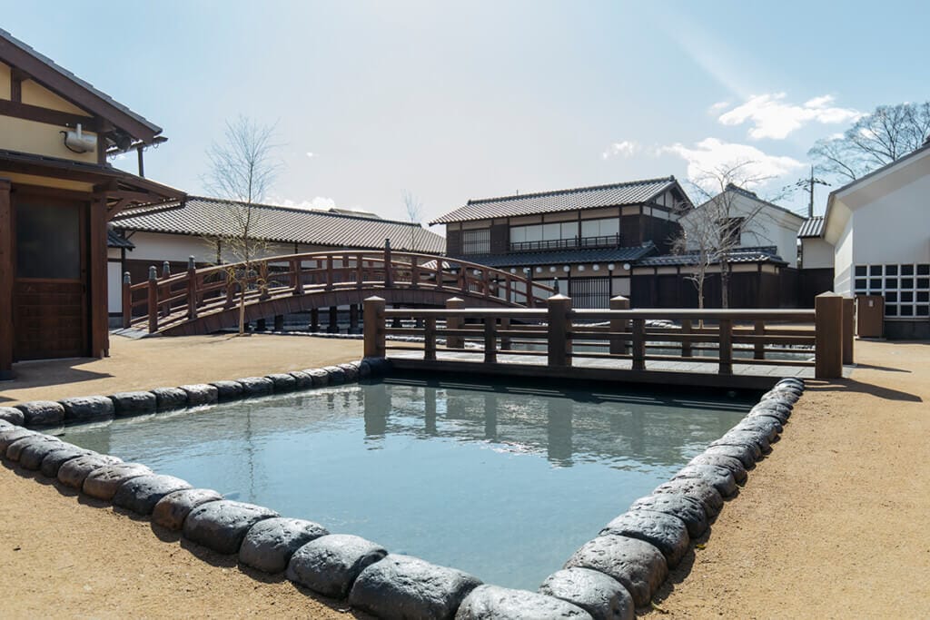 Traditional Japanese courtyard with a wooden arched bridge over a stone-bordered pond and historic buildings in the background.