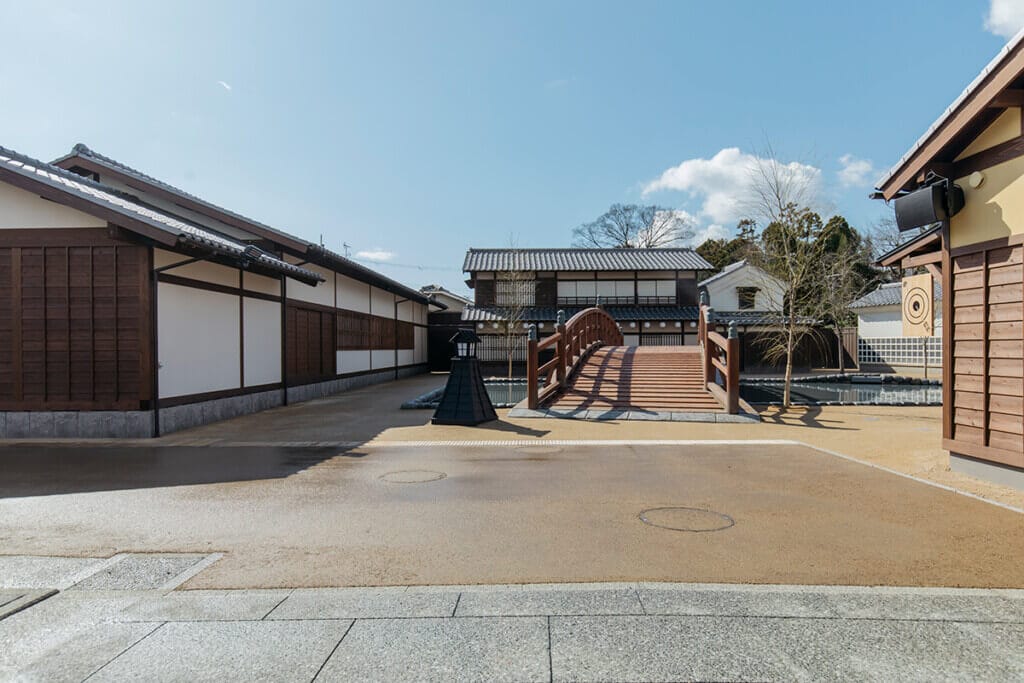 Traditional Japanese courtyard with a wooden arched bridge over a narrow pond, flanked by white-and-brown buildings under a blue sky.