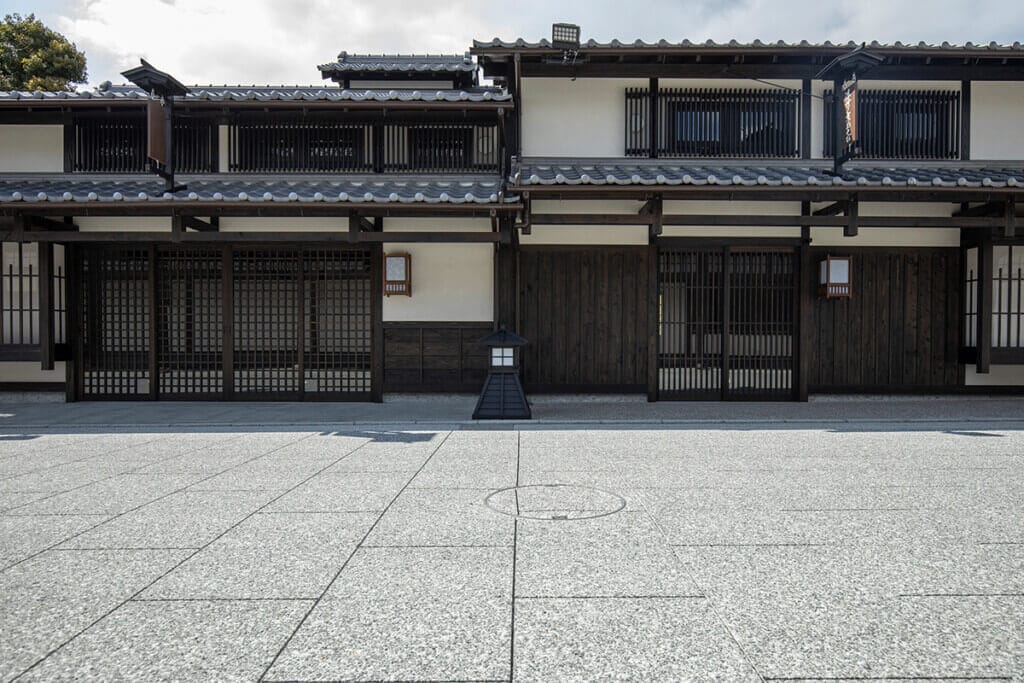 Traditional Japanese wooden building with dark timber framing, white walls, and a stone-paved courtyard in front.