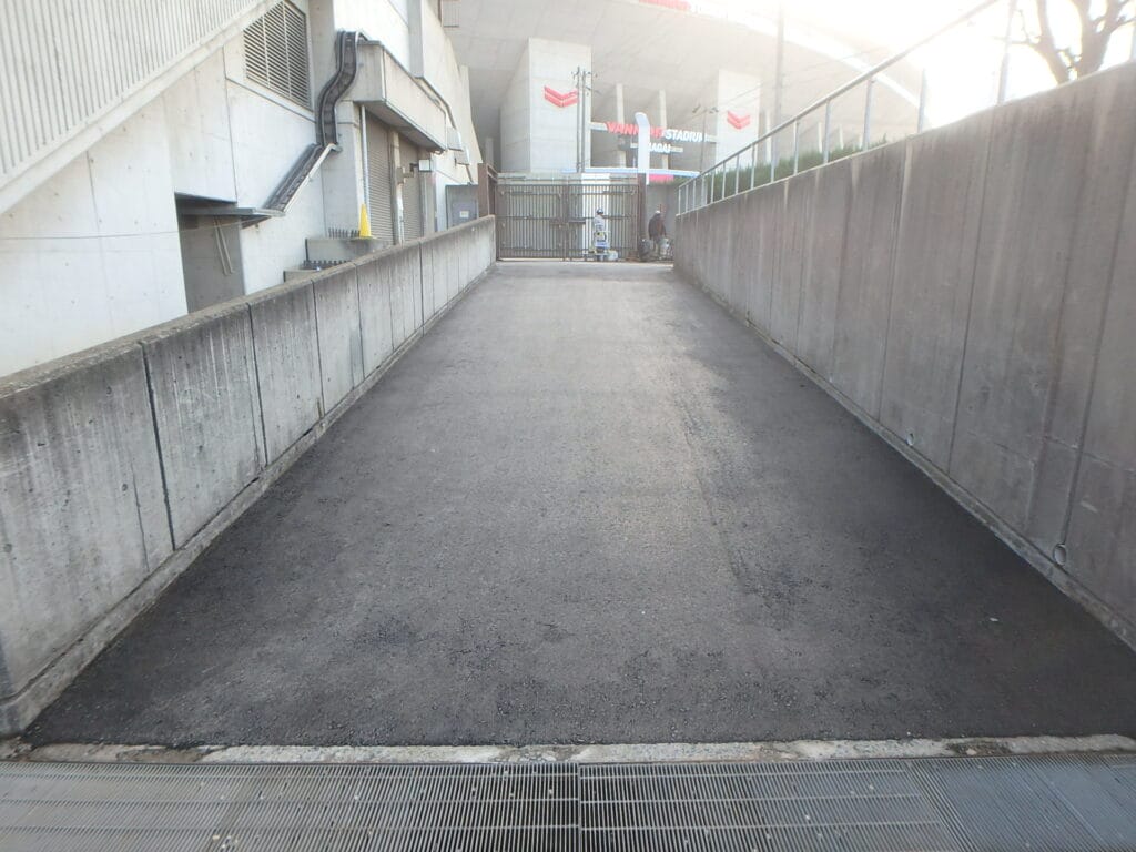 Concrete alley with high concrete walls, metal gate at far end, and a drainage grate in the foreground; two workers near the gate.