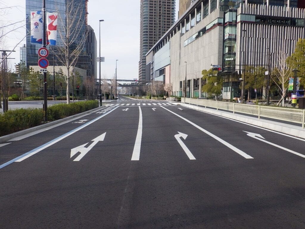 Wide city street with multiple lanes and white directional arrows, modern buildings on both sides, and crosswalk ahead.