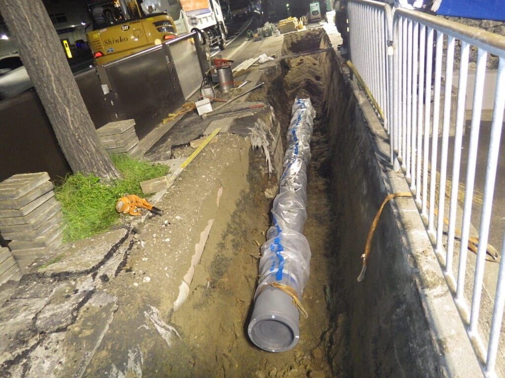 Construction trench along a sidewalk with a long wrapped pipe laid in the ditch and construction tools nearby at night.