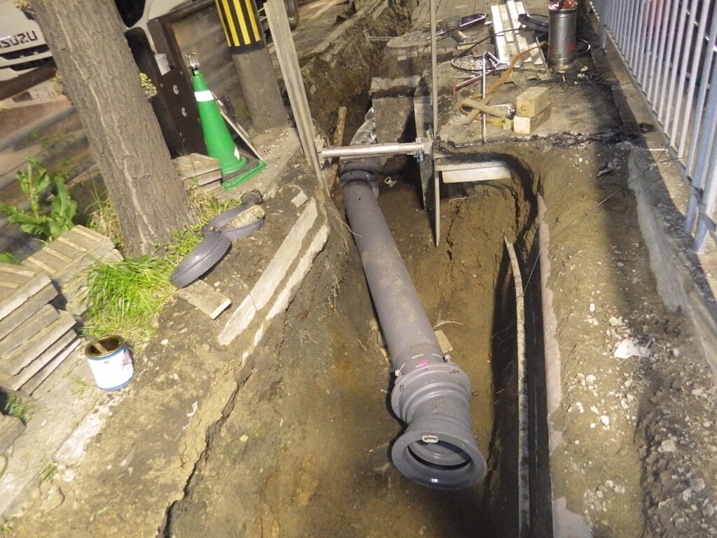 Construction trench with a large gray pipe laid in a dug ditch and equipment scattered nearby, including a green cone and wooden planks.