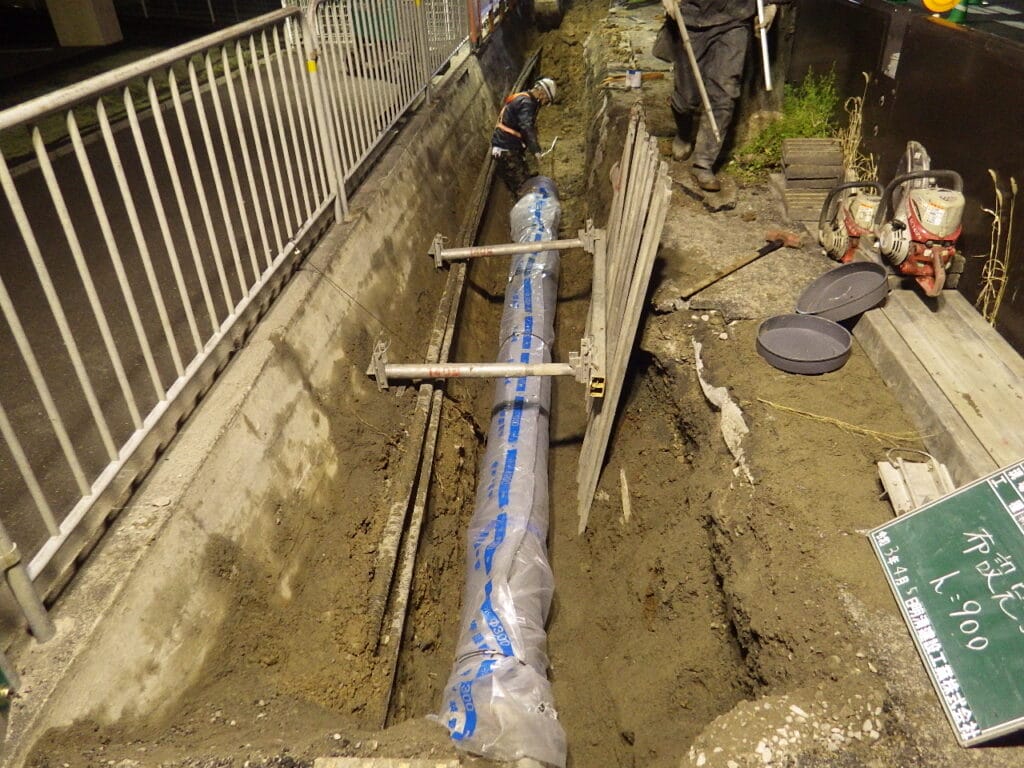 Construction workers install or inspect a buried utility pipe in a trench along a sidewalk, with barriers and tools nearby.