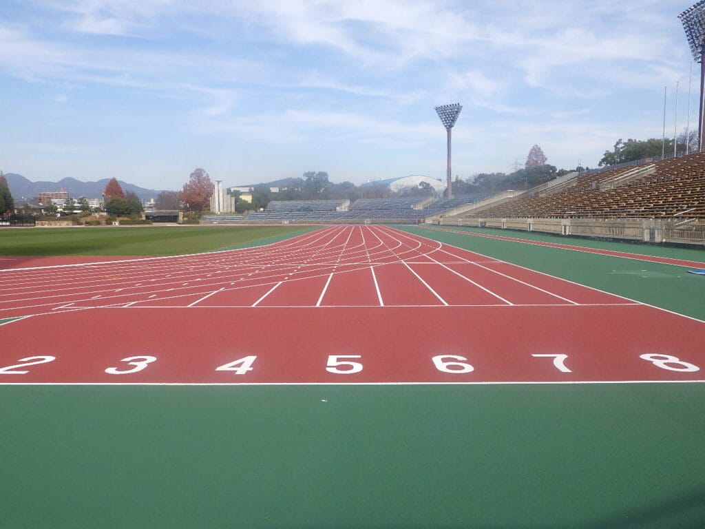 Outdoor track and field stadium with a red running track, white lane markings, and empty grandstands on a sunny day.