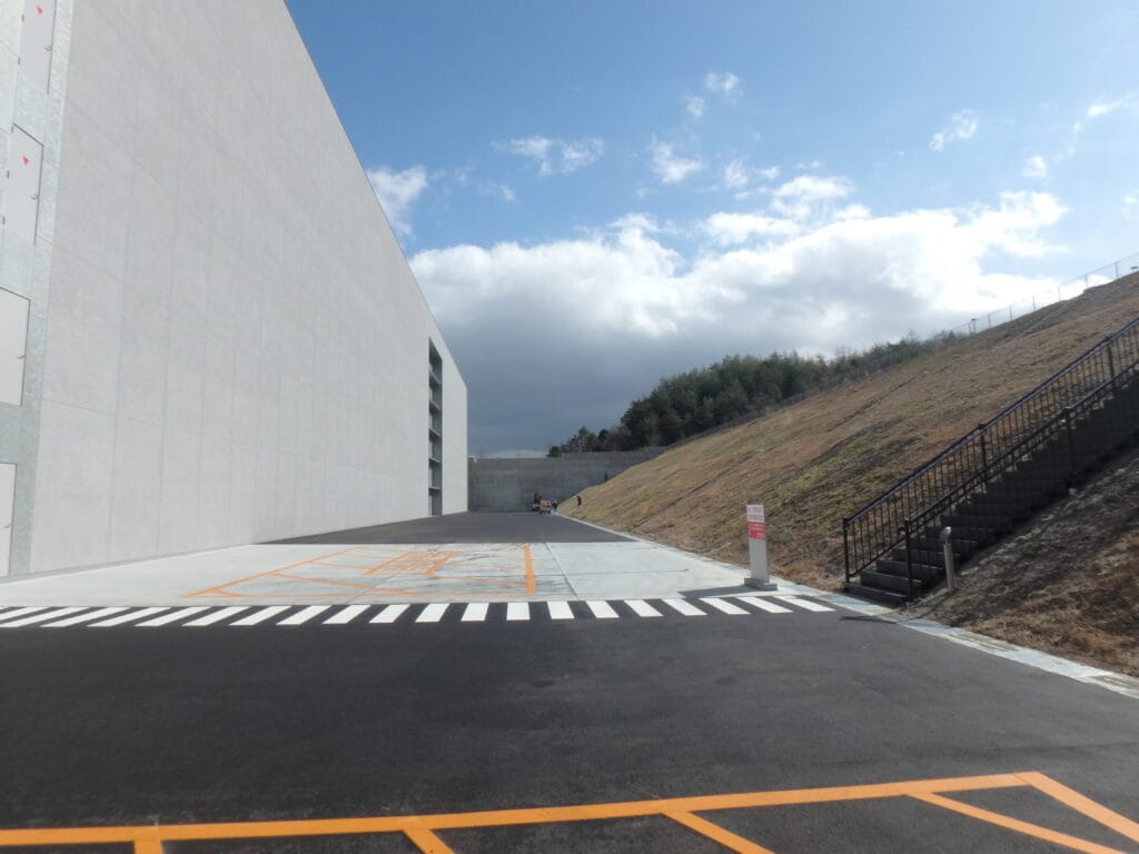 White modern building wall on the left with a sloped grassy hill and metal staircase on the right under a blue sky with clouds.