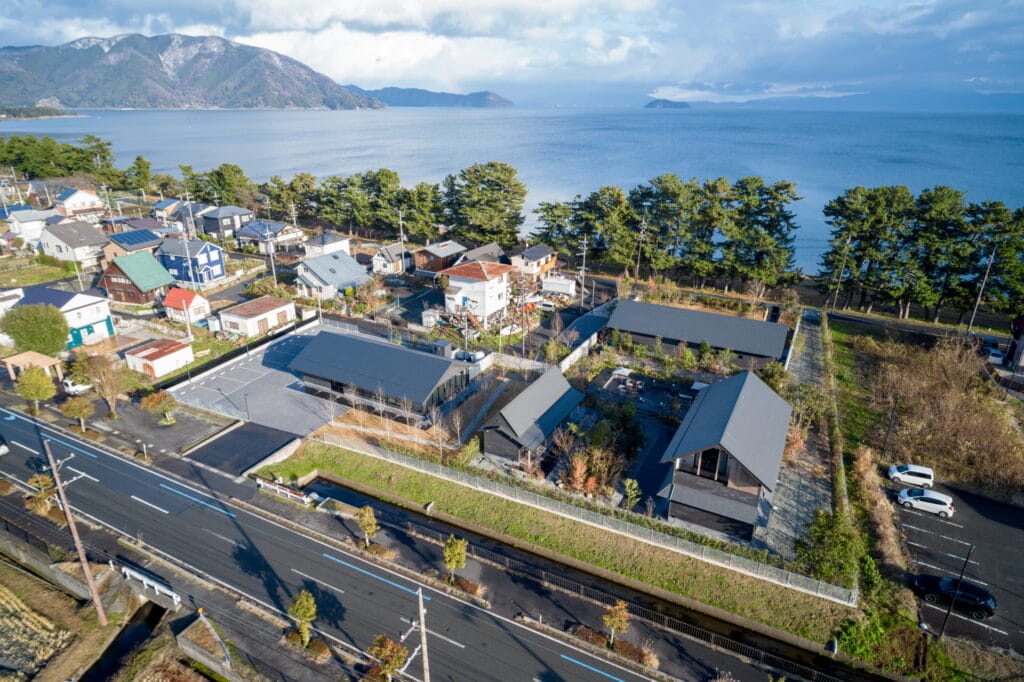 Aerial view of a coastal town with colorful houses along a grid-like street, row of trees by the shore, and calm blue sea with mountains in the distance.