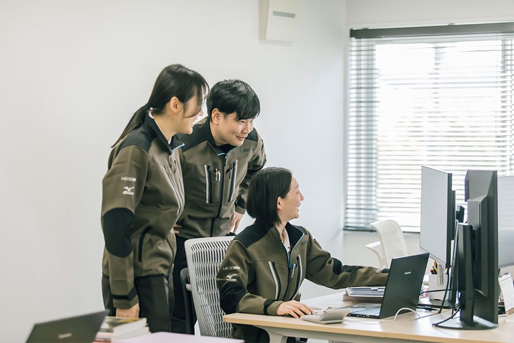Three coworkers in olive uniforms gather around a desk, looking at a computer screen with dual monitors.