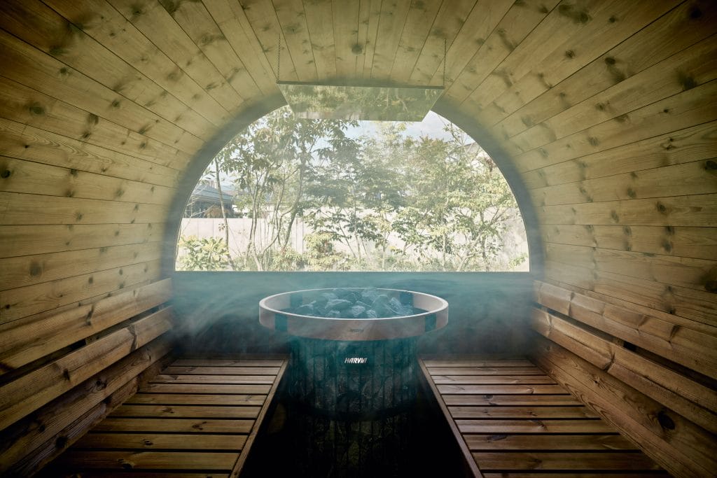 Wooden sauna interior with a circular window, showing greenery outside and a metal heater with stones in the center.