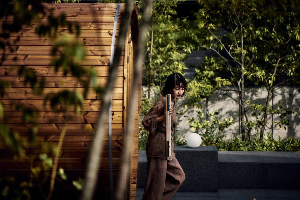 Person standing on outdoor steps beside a wooden wall, holding a vertical metal pole, with greenery in the background.
