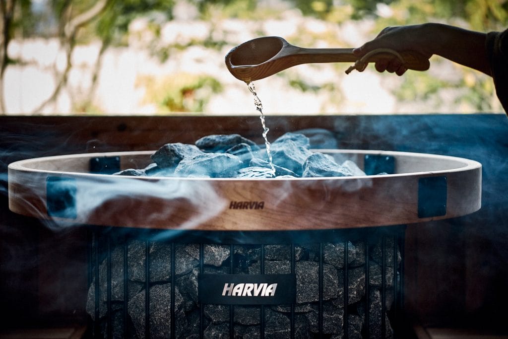 Person pouring water from a wooden scoop onto hot sauna rocks in a Harvia heater.