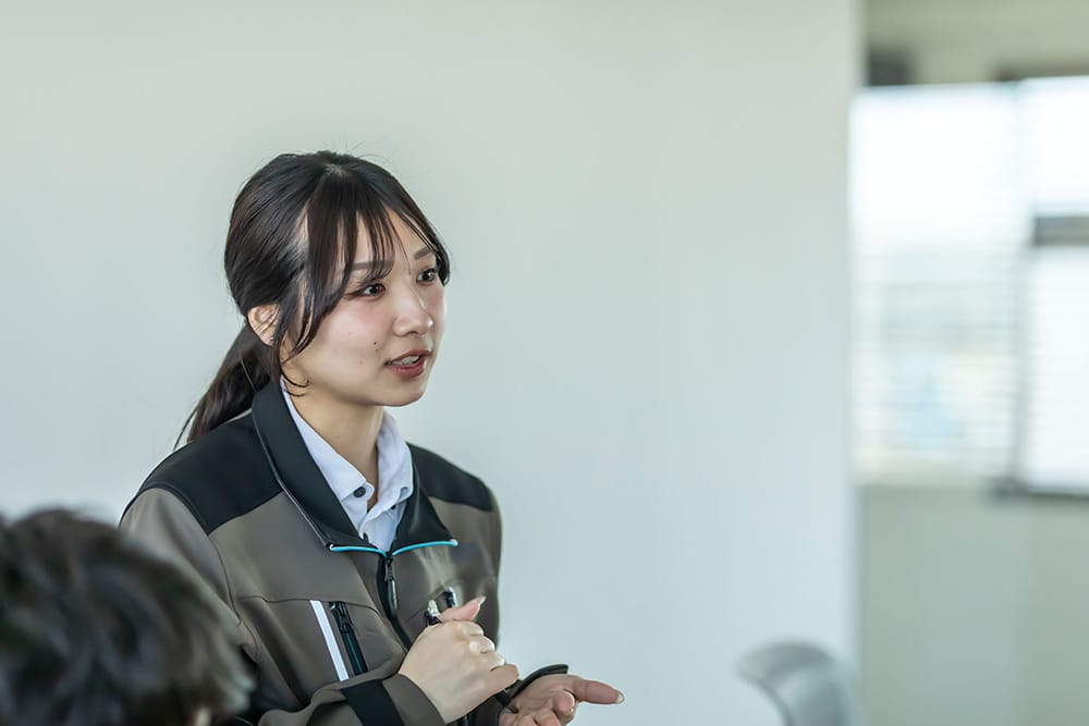 Woman in a gray and black jacket speaks during a meeting, gesturing with her hands.