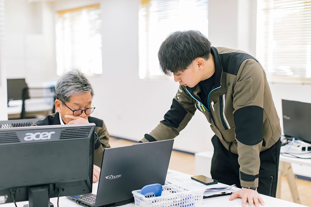 Two men collaborate at a desk—one seated with glasses at an Acer monitor, the other leaning over a laptop, pointing at the screen in a bright office.
