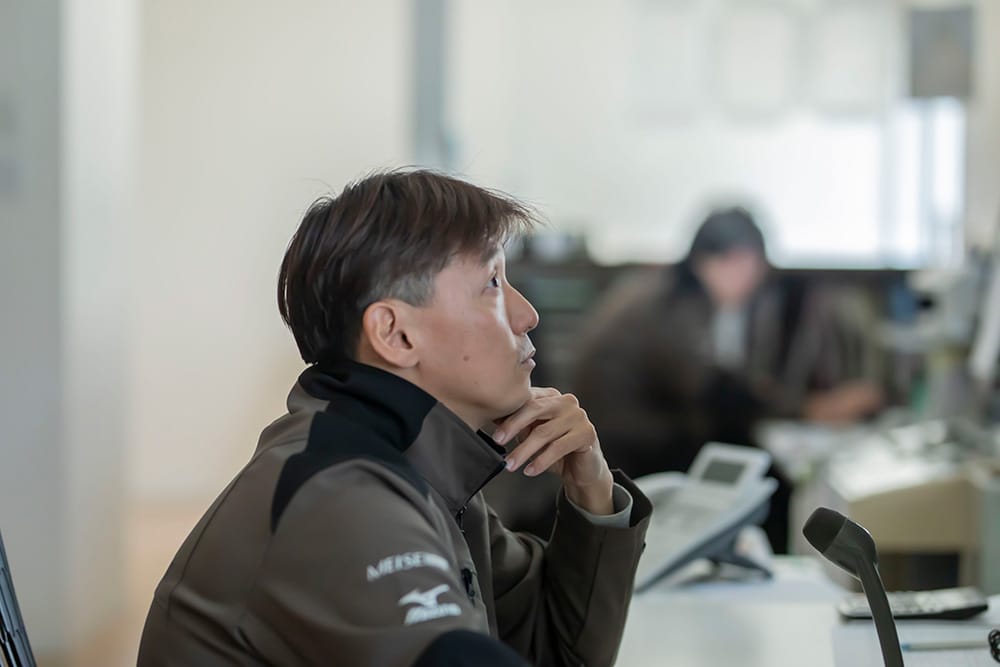 Side profile of a man in a brown jacket resting his chin on his hand at a desk in an office setting.