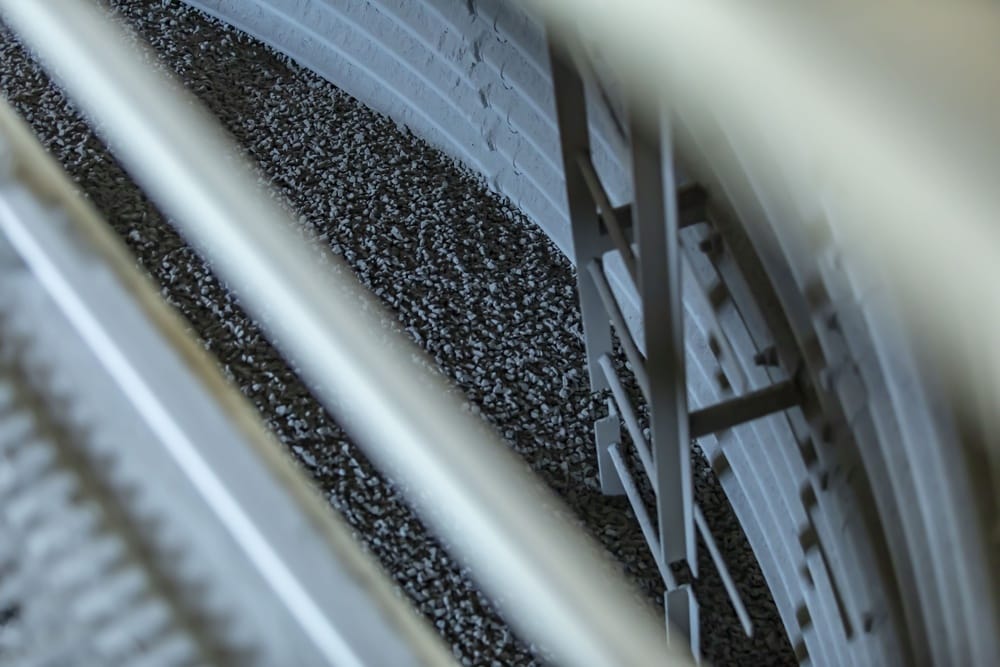 Close-up of a large gray industrial wheel with spokes and a supporting frame, viewed at an angle over a gravel surface.
