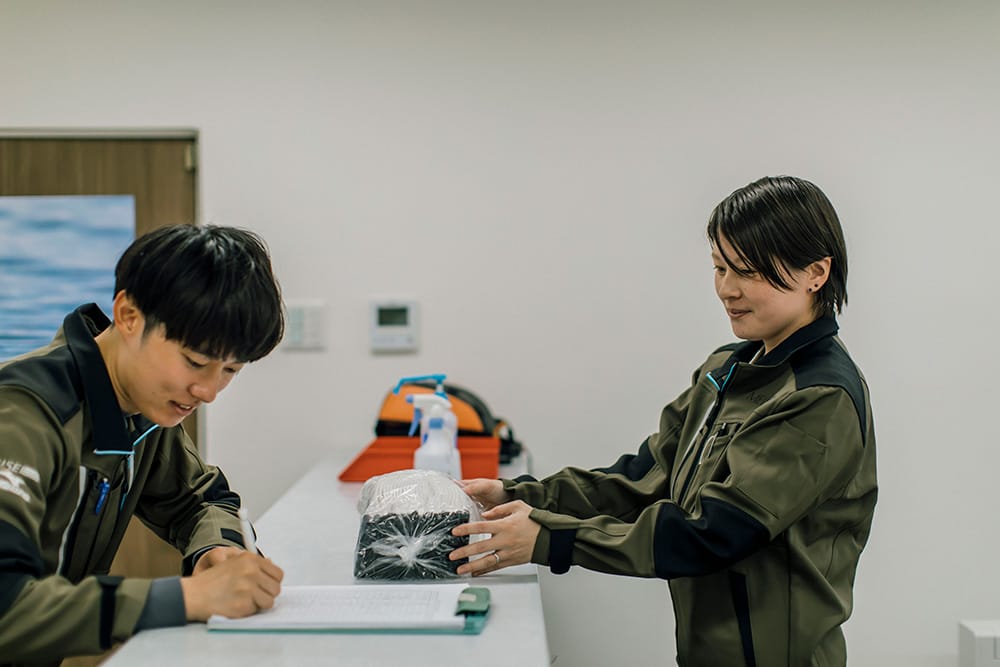 Two people in uniform at a counter as one signs a form while the other hands a wrapped package to them.