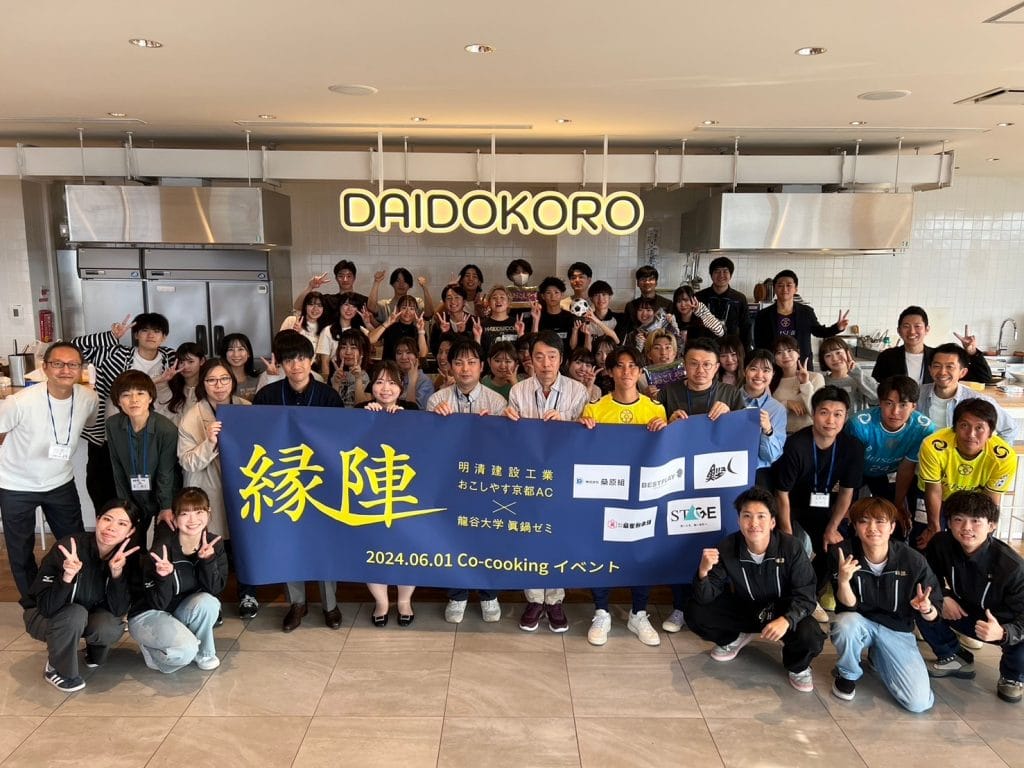 Large group of people posing in a bright kitchen studio, holding a blue banner with yellow Japanese text for a co-cooking event. 'DAIDOKORO' sign visible above them.