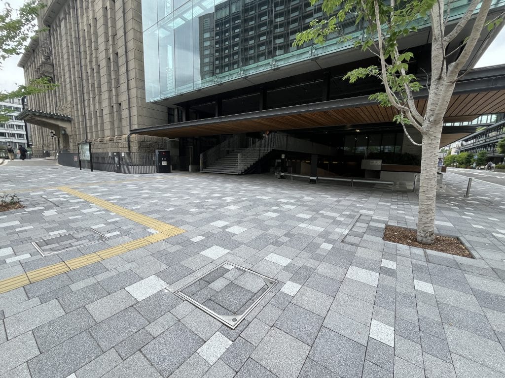Modern urban plaza with a large stone building, glass facade, and a tree in the foreground; tiled pavement and a stairway under a wooden overhang are visible