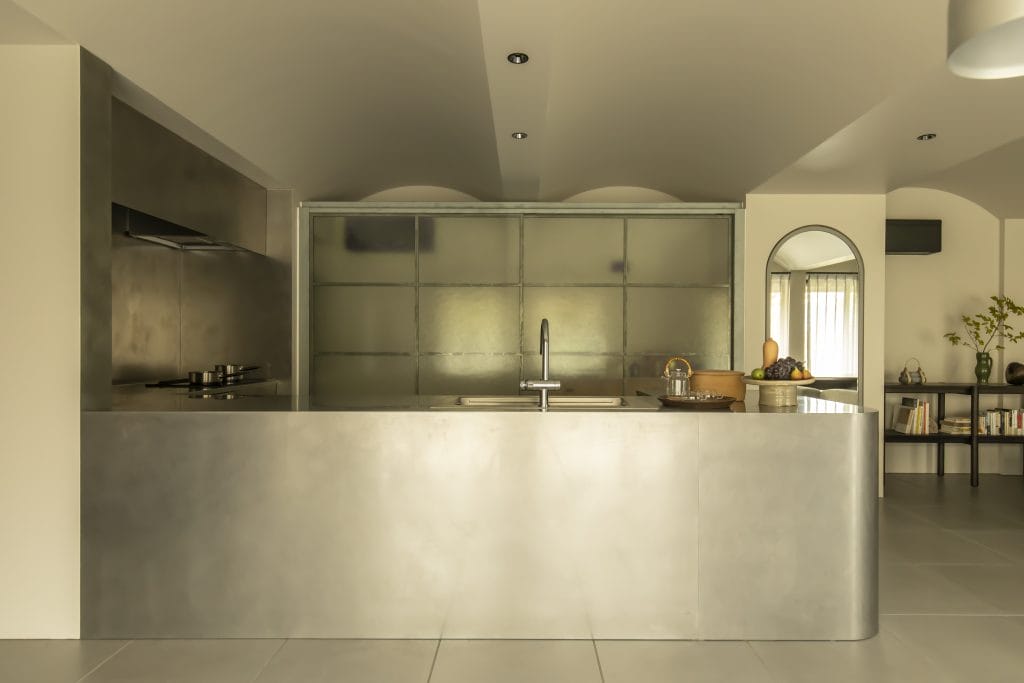 Modern stainless steel kitchen with an island sink and metal backsplash; a round-edged mirror and a fruit bowl visible on the right side.