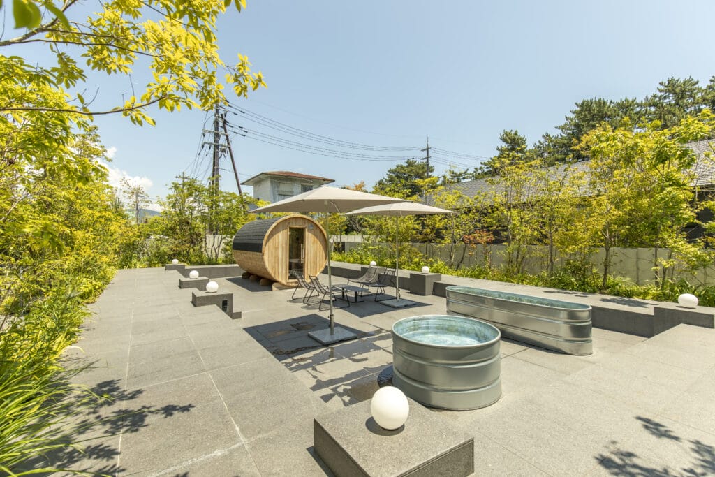 Outdoor patio scene with a round wooden barrel sauna, white umbrellas, and metal chairs amid greenery under a blue sky, bright and serene.