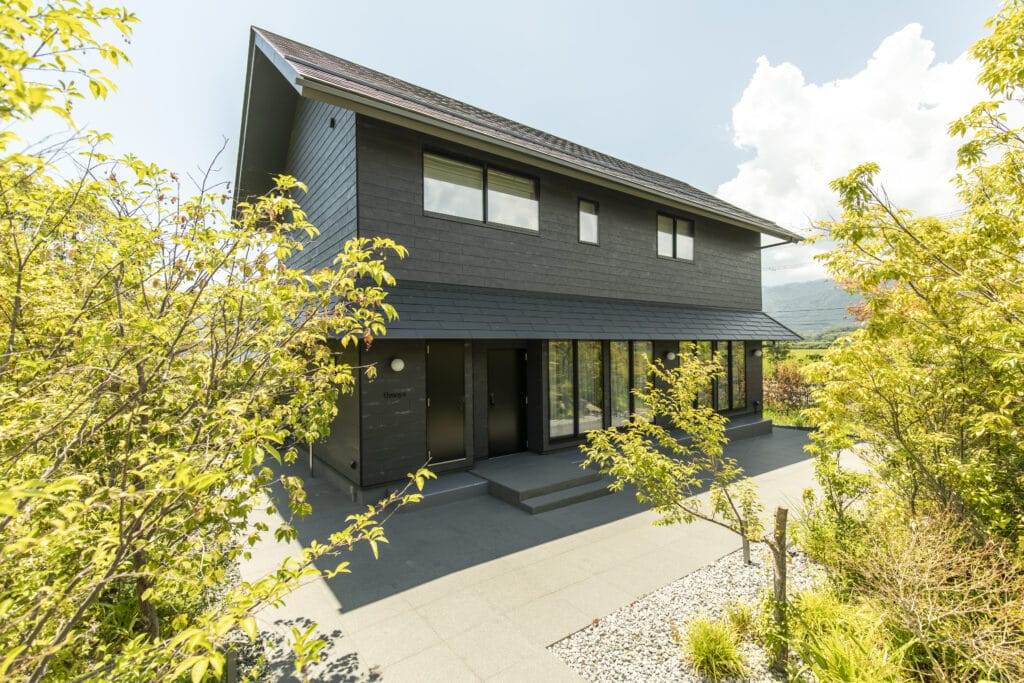 Modern dark gray two-story house with large windows, surrounded by green trees on a sunny day of bright sky.