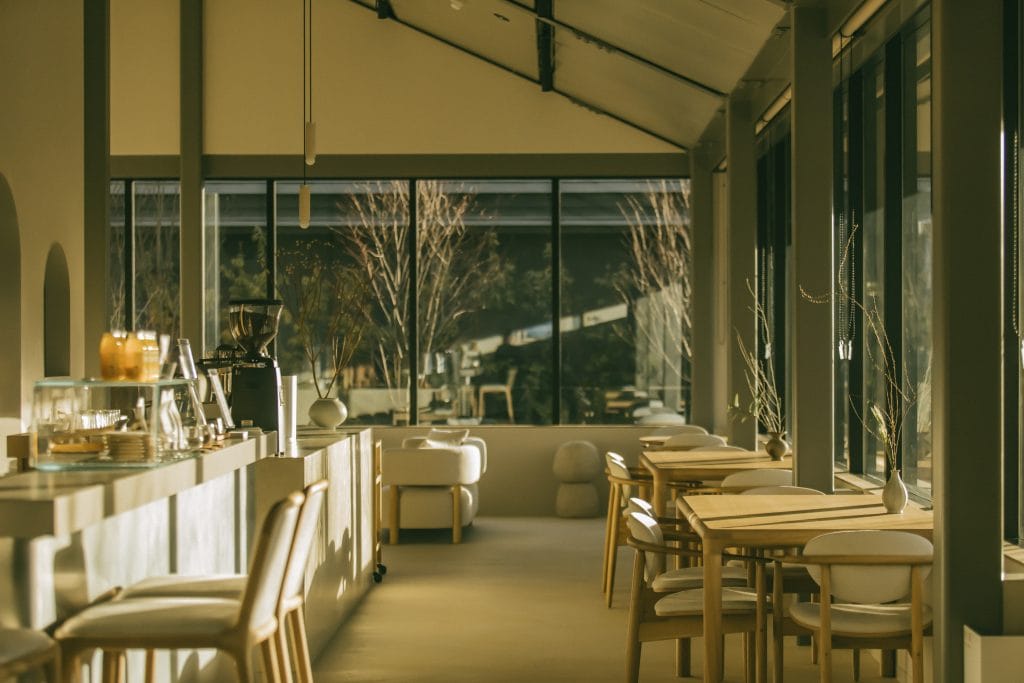 Modern cafe interior with a long light-wood counter on the left and wooden tables and chairs, large windows showing trees outside.
