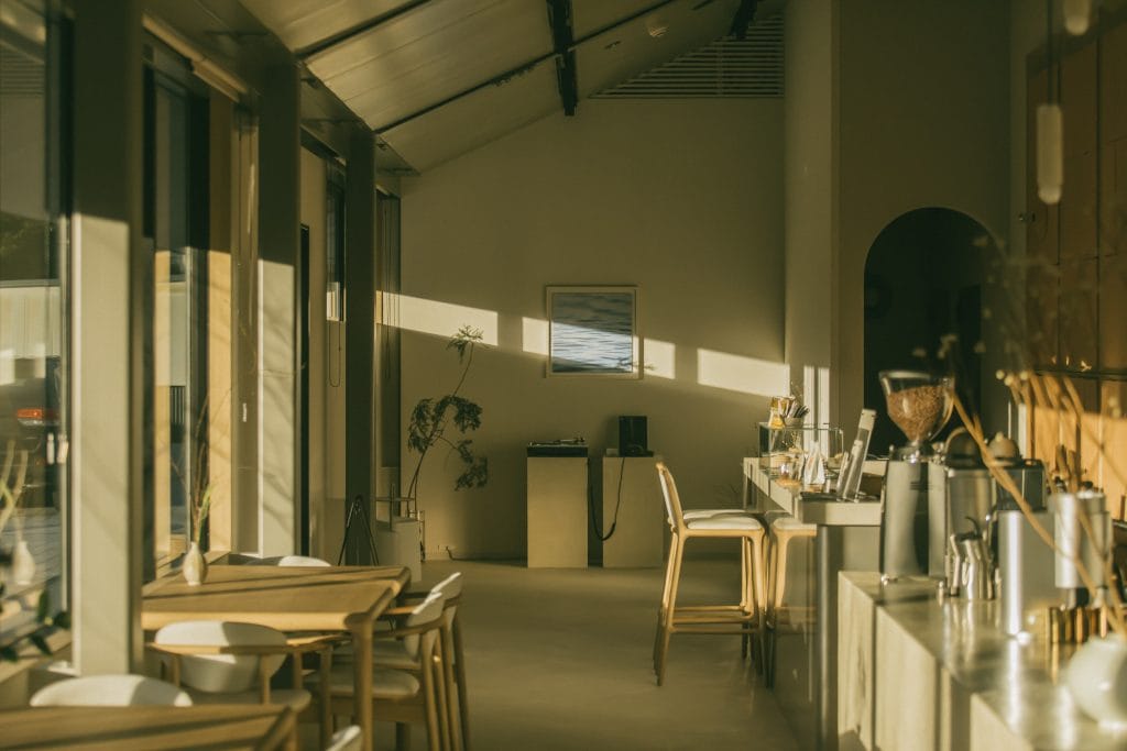 Sunlit cafe interior with wooden tables and chairs along a glass wall, plants near the wall.