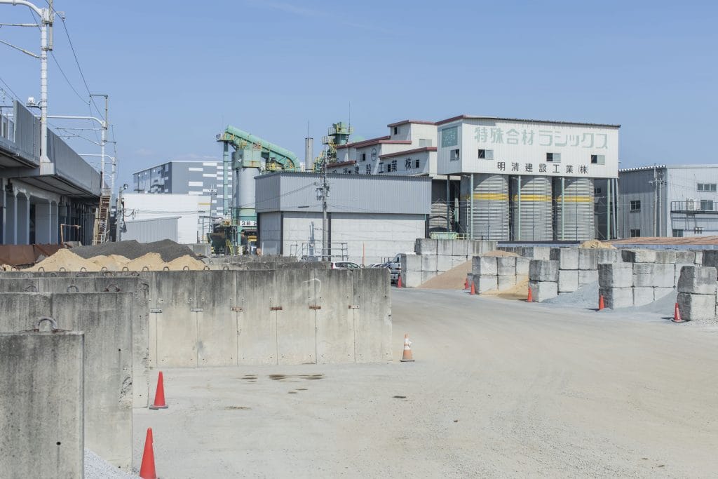 Industrial construction site with concrete barriers, orange safety cones, and a large factory/silo complex under a clear blue sky.