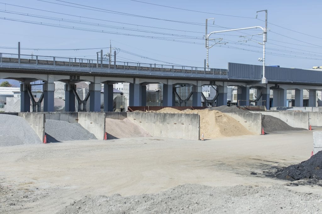 Construction site beneath an elevated roadway with piles of gravel and sand, concrete barriers, and orange cones under a clear blue sky.