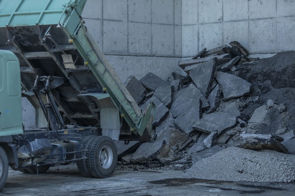 Green industrial conveyor dumping crushed rock and rubble beside a concrete quarry wall, with a pile of broken concrete at its base.
