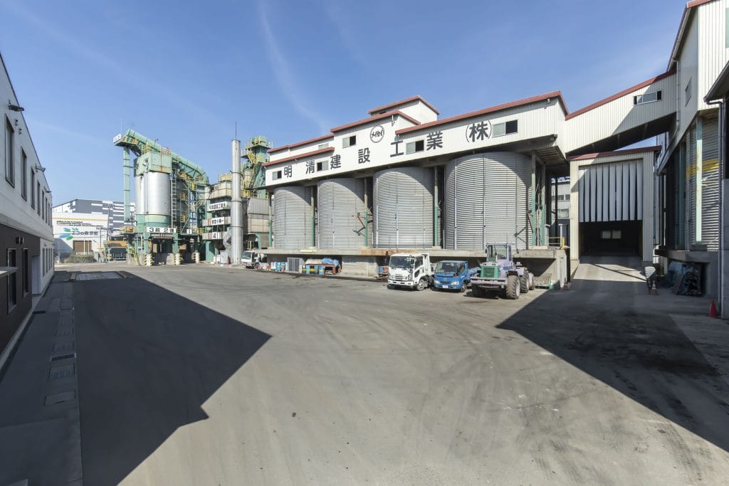 Industrial factory complex with large silos, green machinery, and truck yard under a clear blue sky (Japan signage visible).