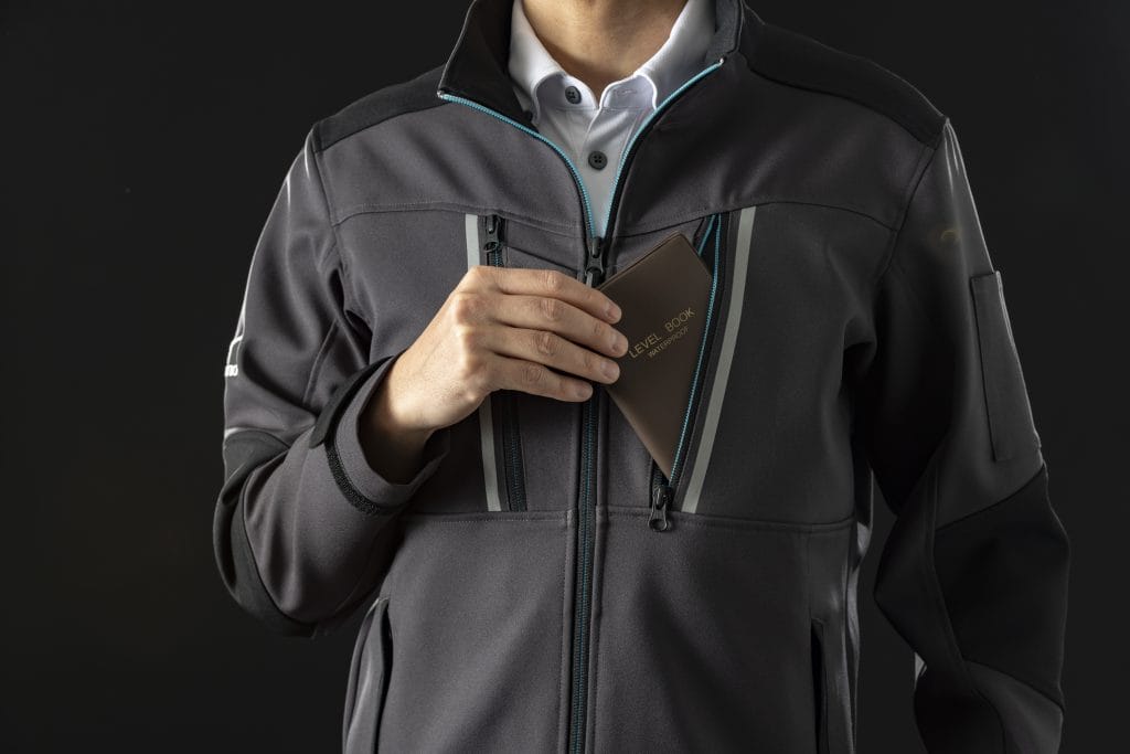 Person in a dark gray waterproof jacket unzipping to reveal a brown passport-style notebook labeled LEVEL BOOK WATERPROOF